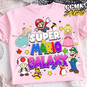 May include: Pink t-shirt featuring the text "SUPER MARIO GALAXY" with colorful cartoon characters. The design includes Mario, Princess Peach, Luigi, and other characters from the Super Mario video game series.