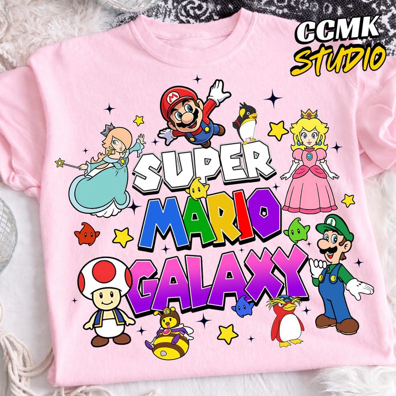 May include: Pink t-shirt featuring the text "SUPER MARIO GALAXY" with colorful cartoon characters. The design includes Mario, Princess Peach, Luigi, and other characters from the Super Mario video game series.