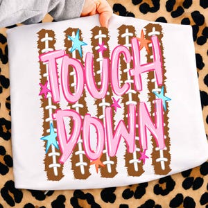 May include: A white t-shirt featuring a graphic print with the text TOUCH DOWN in large pink letters. The design includes vertical brown football-textured stripes with white laces and scattered small blue, pink, and orange stars.