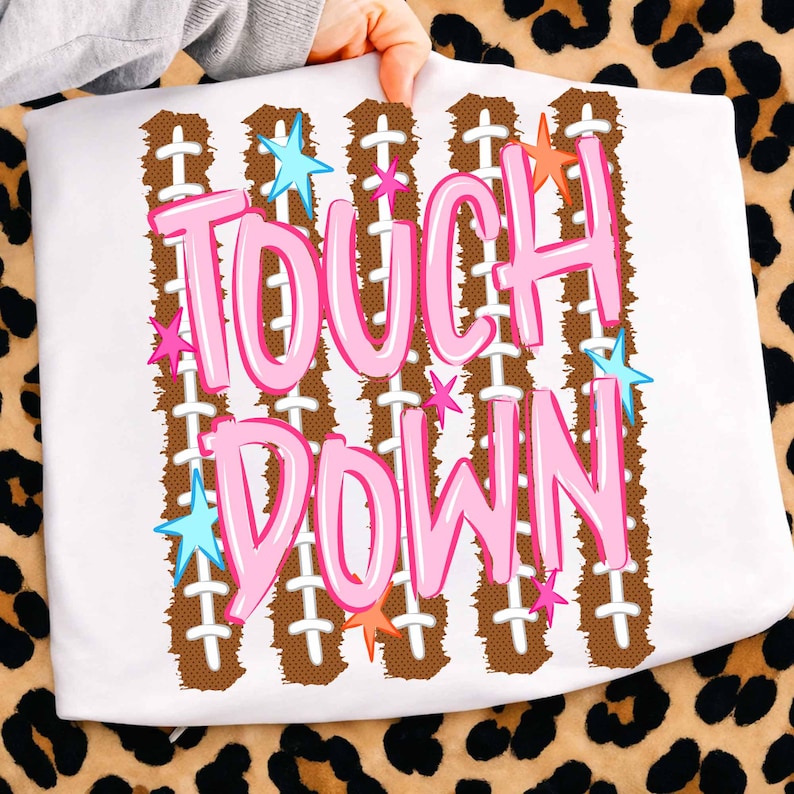 May include: A white t-shirt featuring a graphic print with the text TOUCH DOWN in large pink letters. The design includes vertical brown football-textured stripes with white laces and scattered small blue, pink, and orange stars.