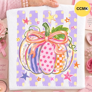 May include: A white fabric square featuring a central pumpkin illustration with a pink bow. The pumpkin has segments with polka dots, checkered patterns, and floral designs. The background has vertical purple stripes with yellow dots and stars.