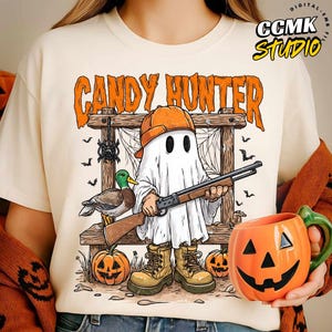 May include: A cream-colored t-shirt featuring a graphic of a ghost wearing an orange cap and tan boots, holding a shotgun. The ghost stands before a wooden frame with a mallard duck, two jack-o'-lanterns, and a spider. The text reads CANDY HUNTER.