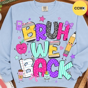 May include: A light blue crewneck sweatshirt featuring a colorful graphic with the text "BRUH WE BACK". The design includes cartoon school supplies like a notebook, apple, pencil, and coffee cup, accented with stars, hearts, and bows in bright colors.