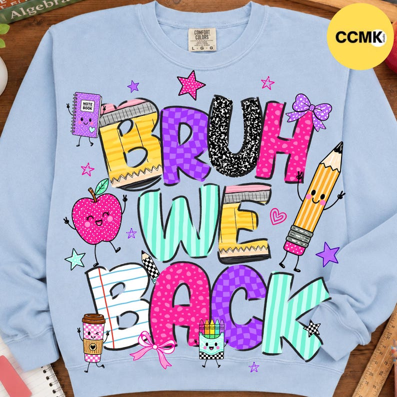 May include: A light blue crewneck sweatshirt featuring a colorful graphic with the text "BRUH WE BACK". The design includes cartoon school supplies like a notebook, apple, pencil, and coffee cup, accented with stars, hearts, and bows in bright colors.
