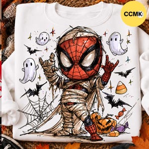 May include: A white sweatshirt featuring a graphic illustration of a character resembling Spider-Man wrapped in mummy bandages. The design includes small white ghosts, black bats, a spider web, orange pumpkins, and candy corn, all rendered in a sketch-like style.