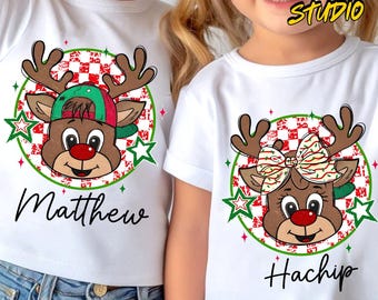 May include: White t-shirts featuring reindeer designs. One reindeer has a green and red cap, the other a bow. The graphics are set within a green circle with stars and a red and white checkered pattern. The names "Matthew" and "Hachip" are written below.