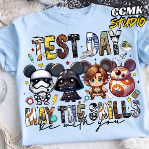 Puede incluir: Camiseta azul claro con las palabras "TEST DAY" y "MAY THE SKILLS be with you". Presenta personajes de dibujos animados, incluyendo Darth Vader, un Stormtrooper y BB-8. La camiseta tiene cuello redondo.