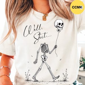 May include: A cream-colored t-shirt featuring a black ink illustration of a skeleton walking while holding a skull-shaped balloon on a string. The text "Well, Shit...." is printed in a black cursive script above the skeleton figure.