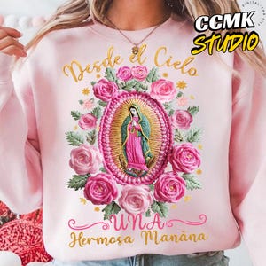 May include: A light pink sweatshirt featuring a detailed embroidered design. The design includes an oval frame with a religious figure, surrounded by pink roses and the text "Desde el Cielo Una Hermosa Mañana" in gold script.