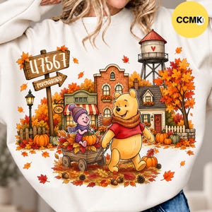 May include: A cream-colored sweatshirt featuring a graphic of Winnie the Pooh and Piglet in an autumn town scene. The design includes a wooden sign reading 47567 Petersburg, a water tower, brick buildings, pumpkins, and falling orange leaves.