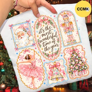 May include: A white t-shirt featuring a festive graphic with the text 'it's the most wonderful time of the year' surrounded by illustrations of Santa Claus, a nutcracker, a ballerina in a pink tutu, a decorated Christmas tree, and a bell.