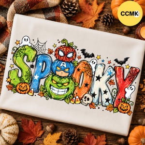 May include: A white t-shirt featuring the word SPOOKY in colorful, stylized letters. The design includes illustrations of pumpkins, ghosts, bats, a spider web, a skull, and candy corn, with superhero-themed pumpkin faces integrated into the text.