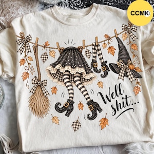 May include: A cream-colored t-shirt featuring a graphic of a witch's skirt, striped stockings, boots, a broom, and a pointed hat hanging on a clothesline with autumn leaves. The text "Well shit..." is printed in black script on the lower right side.