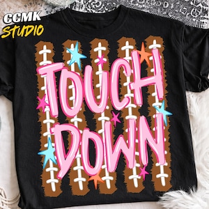 May include: A black t-shirt featuring the text TOUCH DOWN in large pink letters. The background design consists of vertical brown football-textured stripes with white laces, accented by small light blue, pink, and orange star shapes.