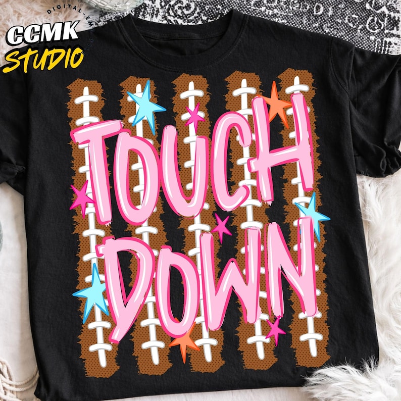 May include: A black t-shirt featuring the text TOUCH DOWN in large pink letters. The background design consists of vertical brown football-textured stripes with white laces, accented by small light blue, pink, and orange star shapes.