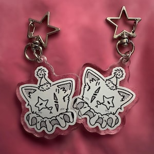 May include: Two clear acrylic keyrings with a pink border, each featuring a black and white cartoon cat design wearing a party hat. The keyrings have silver star-shaped clasps.