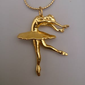 May include: A gold chain necklace with a pendant of a ballerina in a gold dress. The ballerina is in a dancing pose with her arms outstretched.