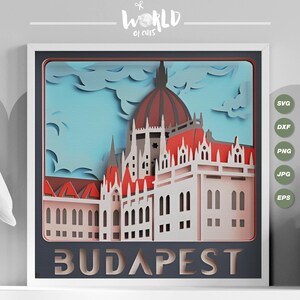 May include: A paper cut-out design of the Hungarian Parliament Building in Budapest. The building is depicted in a 3D style with a red roof and white walls. The design is framed in a white frame with a black background. The text "BUDAPEST" is displayed at the bottom of the design.