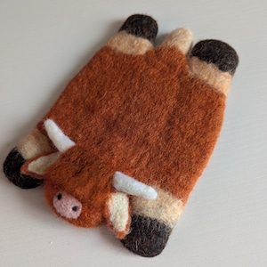 May include: Two felted wool coasters in the shape of a Highland cow. The coasters are predominantly brown with black hooves and white horns. The cow's face has a pink nose and small black eyes. These handmade coasters add a rustic touch to any home.