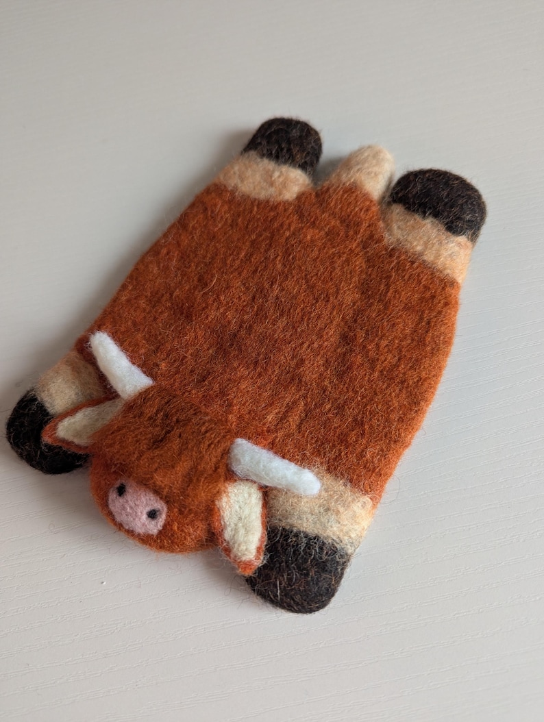 May include: Two felted wool coasters in the shape of a Highland cow. The coasters are predominantly brown with black hooves and white horns. The cow's face has a pink nose and small black eyes. These handmade coasters add a rustic touch to any home.