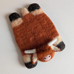 May include: A handcrafted felt Highland cow-shaped item. The cow is mainly brown with black hooves and horns, and a pink nose. The felted item is designed to resemble a Highland cow.
