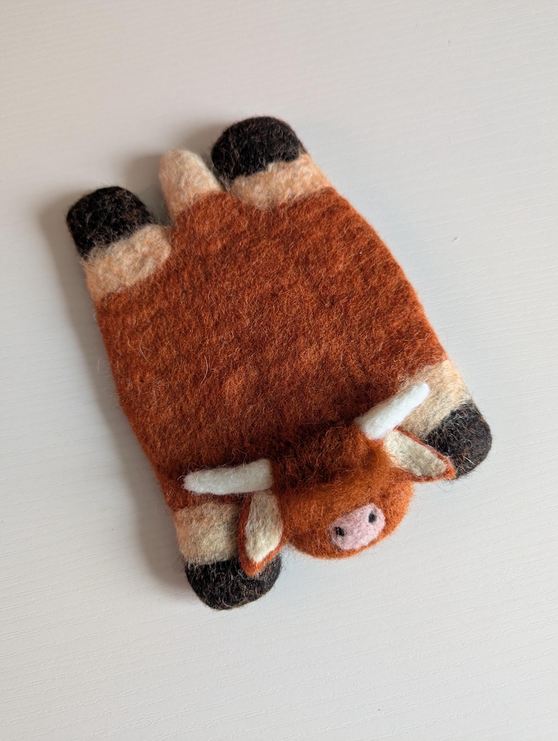 May include: A handcrafted felt Highland cow-shaped item. The cow is mainly brown with black hooves and horns, and a pink nose. The felted item is designed to resemble a Highland cow.