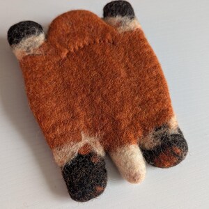 May include: A handcrafted felt fox-shaped item in shades of orange, black, and cream. The fox design has three finger-like shapes with black tips and cream accents. This item is likely a decorative piece or a cosy accessory.