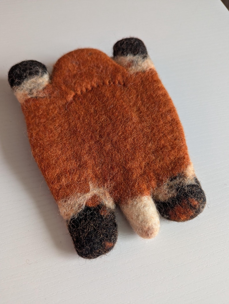 May include: A handcrafted felt fox-shaped item in shades of orange, black, and cream. The fox design has three finger-like shapes with black tips and cream accents. This item is likely a decorative piece or a cosy accessory.