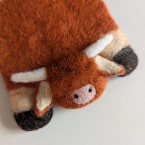 May include: A handcrafted felt Highland cow decoration. The ornament is mainly orange-brown with white horns and details. The cow's face features a pink nose and small black eyes. The felted texture gives it a soft, handmade look.