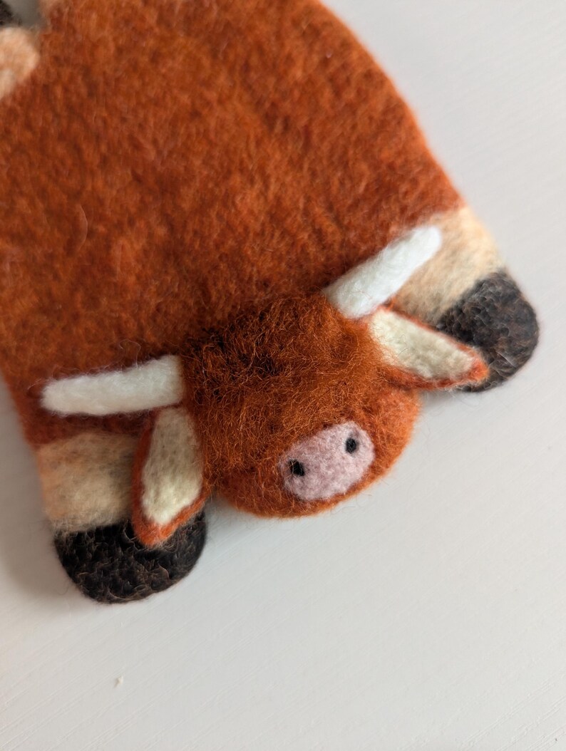 May include: A handcrafted felt Highland cow decoration. The ornament is mainly orange-brown with white horns and details. The cow's face features a pink nose and small black eyes. The felted texture gives it a soft, handmade look.