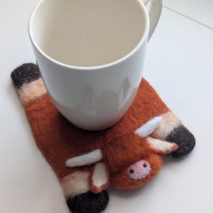 May include: A white ceramic mug sits on a handmade felt coaster shaped like a brown cow. The coaster has white horns, a pink nose, and black and tan hooves. The mug has a curved handle.
