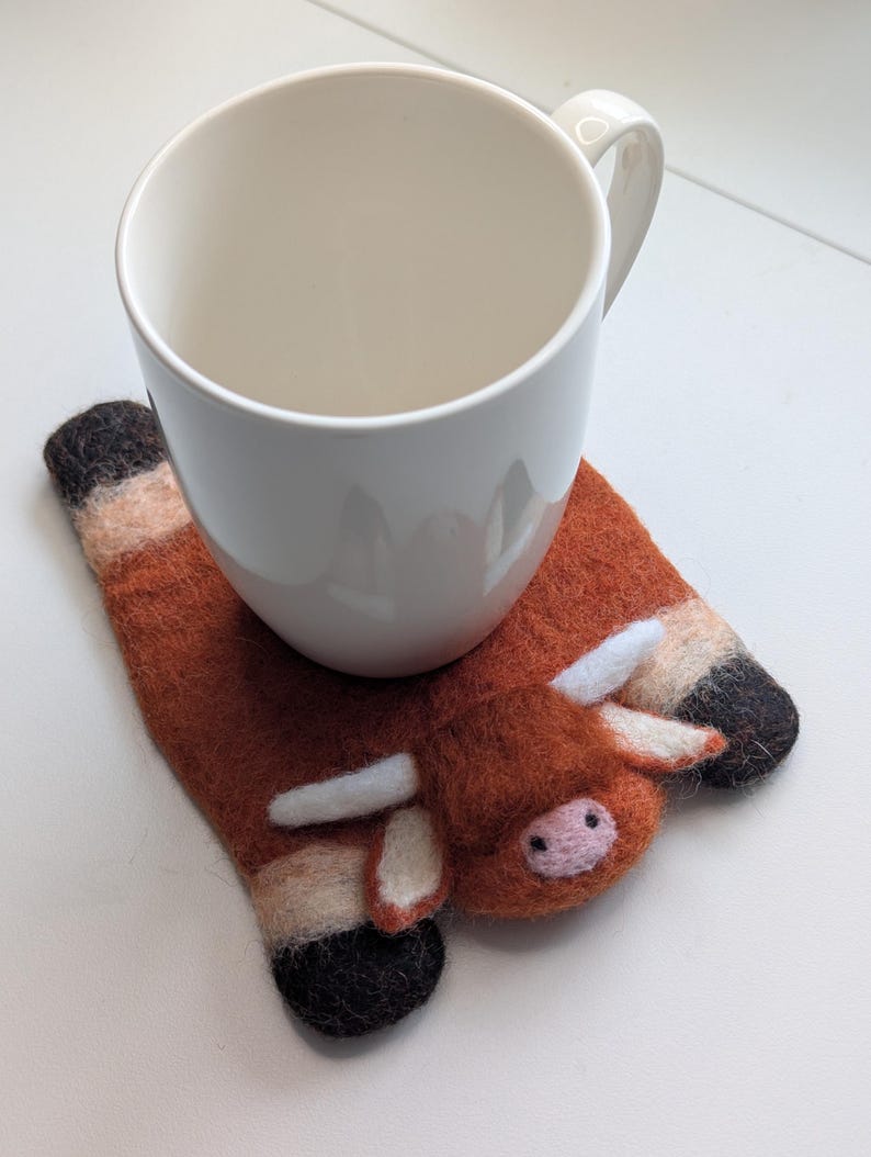 May include: A white ceramic mug sits on a handmade felt coaster shaped like a brown cow. The coaster has white horns, a pink nose, and black and tan hooves. The mug has a curved handle.
