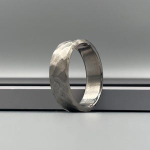 May include: A silver ring with a hammered texture. The band-style ring has a textured surface. The ring is displayed against a grey background. The ring's design is simple yet striking.