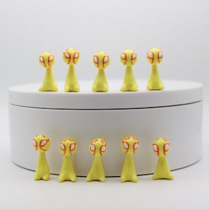 Pode incluir: Uma coleção de dez estatuetas amarelas com um design único. Cada estatueta tem uma cabeça arredondada com detalhes laranja e brancos, sobre um corpo fino e amarelo. Estão dispostas numa plataforma branca e circular contra um fundo branco liso.