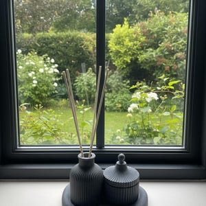 May include: A dark grey ceramic diffuser set with a lidded jar and a tray. The diffuser has reeds inserted. The set is placed in front of a window overlooking a green garden. The objects are on a white surface.