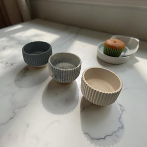 May include: Three small, ribbed, ceramic bowls in shades of grey and beige are arranged on a white marble surface. A small white dish with a cupcake is also present. The bowls have a minimalist design.