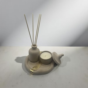 May include: A beige decorative set comprising a small vase with reeds, a candle in a lidded container, and a round tray with gold leaf accents. The items are arranged on a white surface, creating a minimalist aesthetic.