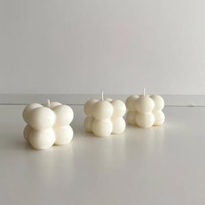 May include: Three ivory-coloured, cube-shaped candles with a bubble design. Each candle is composed of rounded spheres, with a single wick at the top. The candles are arranged on a white surface against a plain white background.