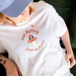Può includere: Maglietta bianca con un'immagine di un fungo rosso e il testo "not flirting just autistic".