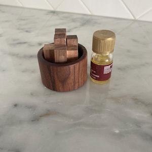 May include: A wooden puzzle with small, rectangular blocks in a round wooden container. Beside it is a small glass bottle with a gold cap and a brown label. The bottle contains a yellow liquid.