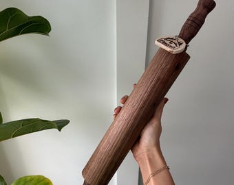 May include: A wooden rolling pin with a brown wood finish and a small wooden tag attached to the handle. The tag has the word "Handcraft" written on it.