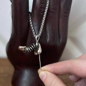 Peut inclure: Un collier pendentif argenté avec un design abstrait unique. Le pendentif a une forme de spirale et de goutte d'eau. La chaîne du collier est de style gourmette. Le bijou est présenté sur une main en bois sombre.
