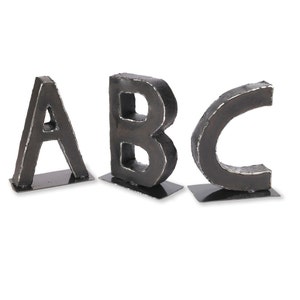 May include: Three metal letters, A, B, and C, are standing on individual bases. The letters are black and have a rough, textured surface.