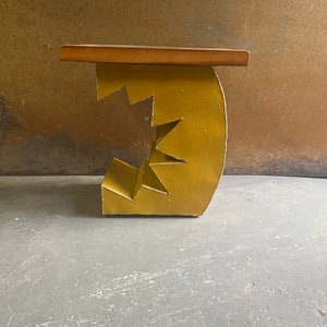 K&ouml;nnte beinhalten: Ein gelber Holzhocker mit einem einzigartigen, geometrischen Design. Der Hocker hat eine flache, rechteckige Oberseite und eine Basis, die einem Zahnrad oder einer Reihe ineinandergreifender Formen &auml;hnelt.