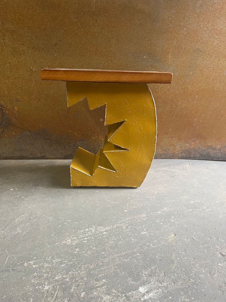 K&ouml;nnte beinhalten: Ein gelber Holzhocker mit einem einzigartigen, geometrischen Design. Der Hocker hat eine flache, rechteckige Oberseite und eine Basis, die einem Zahnrad oder einer Reihe ineinandergreifender Formen &auml;hnelt.