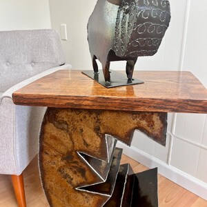 K&ouml;nnte beinhalten: Ein rustikaler Holztisch mit einem Metallgestell mit geometrischem Design. Der Tisch hat eine dunkelbraune Holzplatte und ein Metallgestell mit einem einzigartigen, abstrakten Design. Eine Metallsculptur eines Stiers mit einem detaillierten Design steht auf dem Tisch.