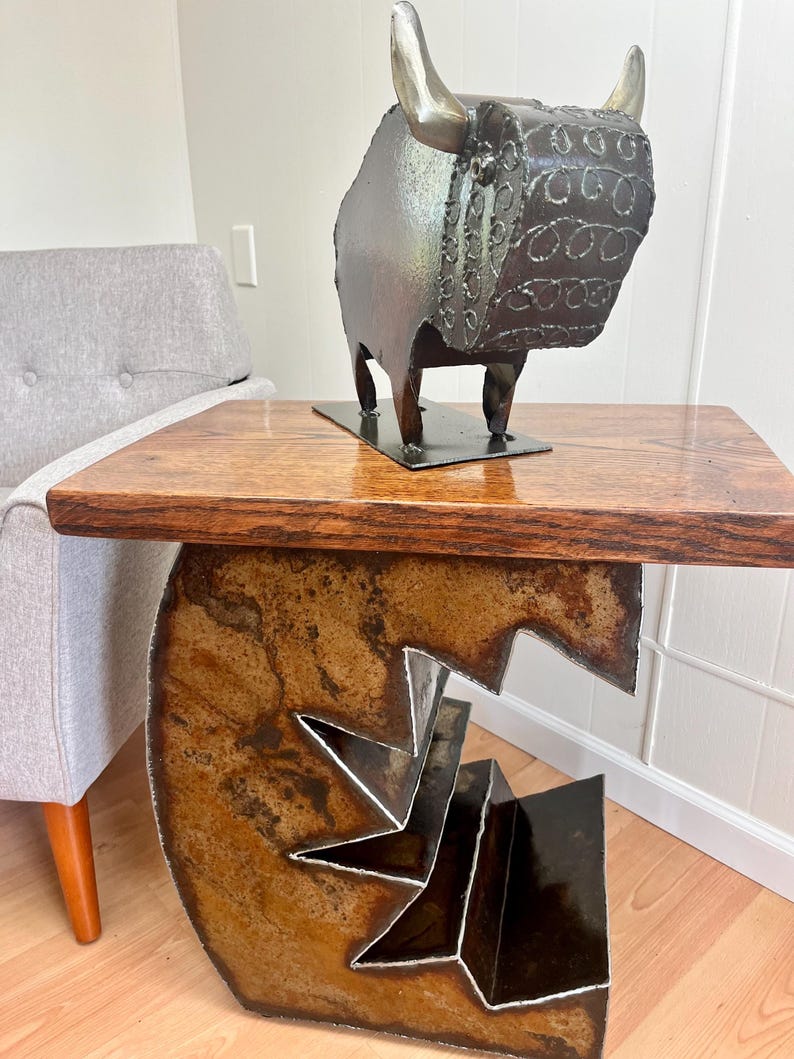 K&ouml;nnte beinhalten: Ein rustikaler Holztisch mit einem Metallgestell mit geometrischem Design. Der Tisch hat eine dunkelbraune Holzplatte und ein Metallgestell mit einem einzigartigen, abstrakten Design. Eine Metallsculptur eines Stiers mit einem detaillierten Design steht auf dem Tisch.