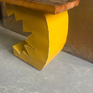 K&ouml;nnte beinhalten: Eine gelbe Holzbank mit einem einzigartigen, abstrakten Design. Die Bank hat eine geschwungene R&uuml;ckenlehne und eine flache Sitzfl&auml;che. Die Bank ist aus Holz und gelb lackiert.