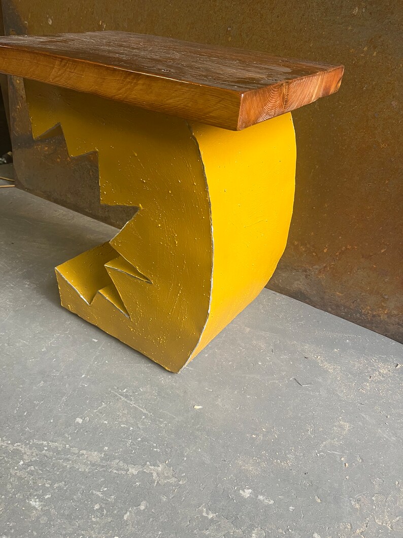 K&ouml;nnte beinhalten: Eine gelbe Holzbank mit einem einzigartigen, abstrakten Design. Die Bank hat eine geschwungene R&uuml;ckenlehne und eine flache Sitzfl&auml;che. Die Bank ist aus Holz und gelb lackiert.