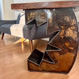 K&ouml;nnte beinhalten: Eine rustikale Holztischplatte mit einem einzigartigen, abstrakten Metallfu&szlig;. Der Fu&szlig; besteht aus mehreren zusammengeschwei&szlig;ten Metallteilen und hat eine rostige Oberfl&auml;che.